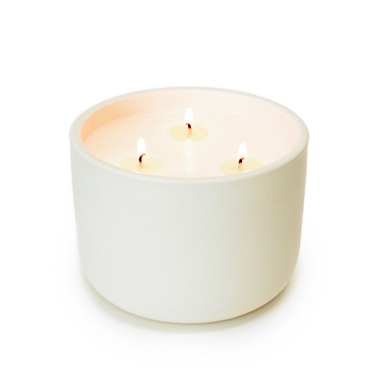 A lit white ceramic candle holder with three wicks, emitting a warm glow, set against a seamless white background.